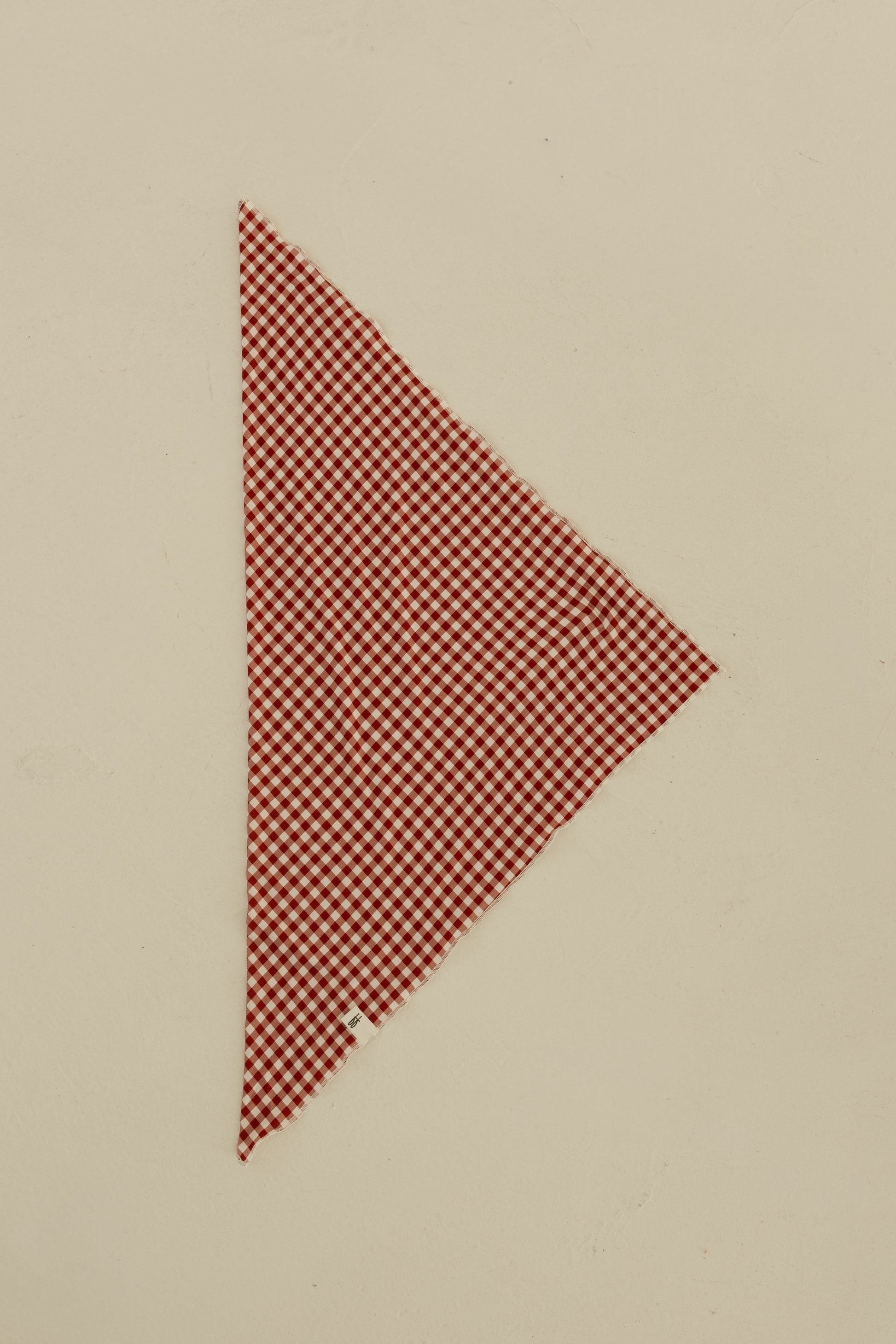 The Bamboo Bandana | Red Gingham by forever french baby, cut in a right triangle shape, is displayed on a plain beige surface.