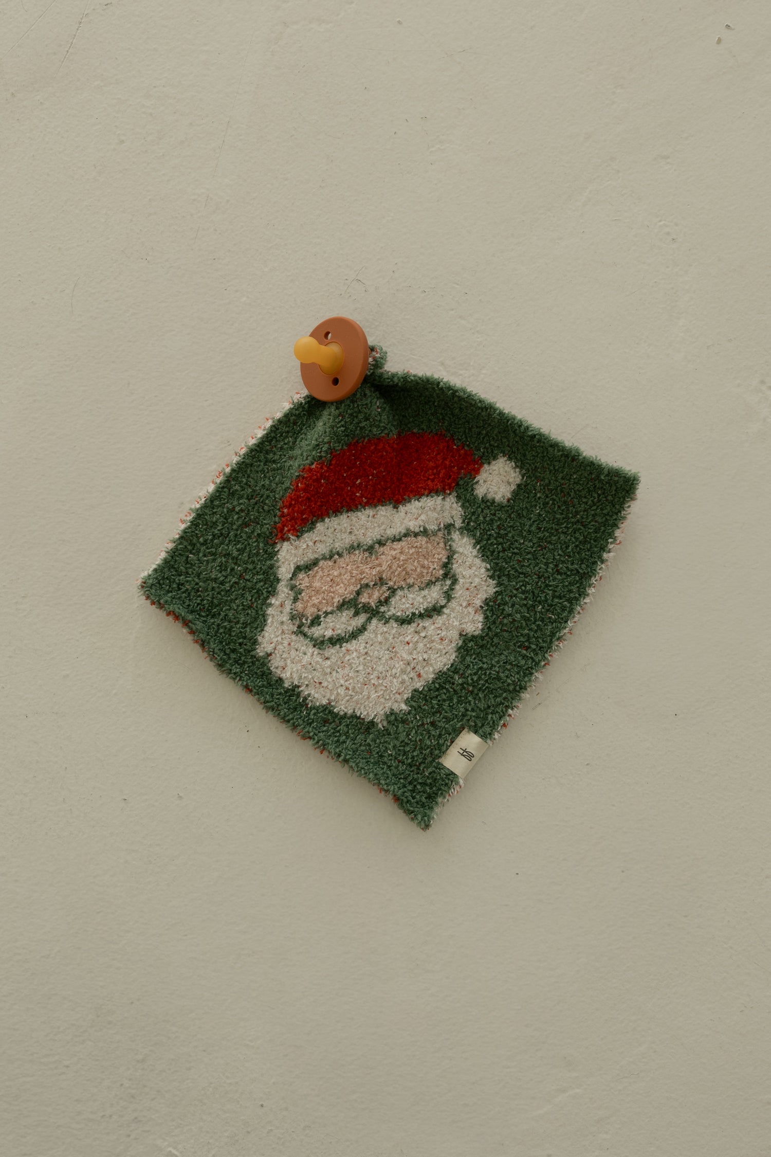 The Lovey | Santa Checkerboard by forever french baby, featuring a green square cloth with a Santa face in a red hat, is pinned to a beige wall with a round brown bird-shaped push pin.