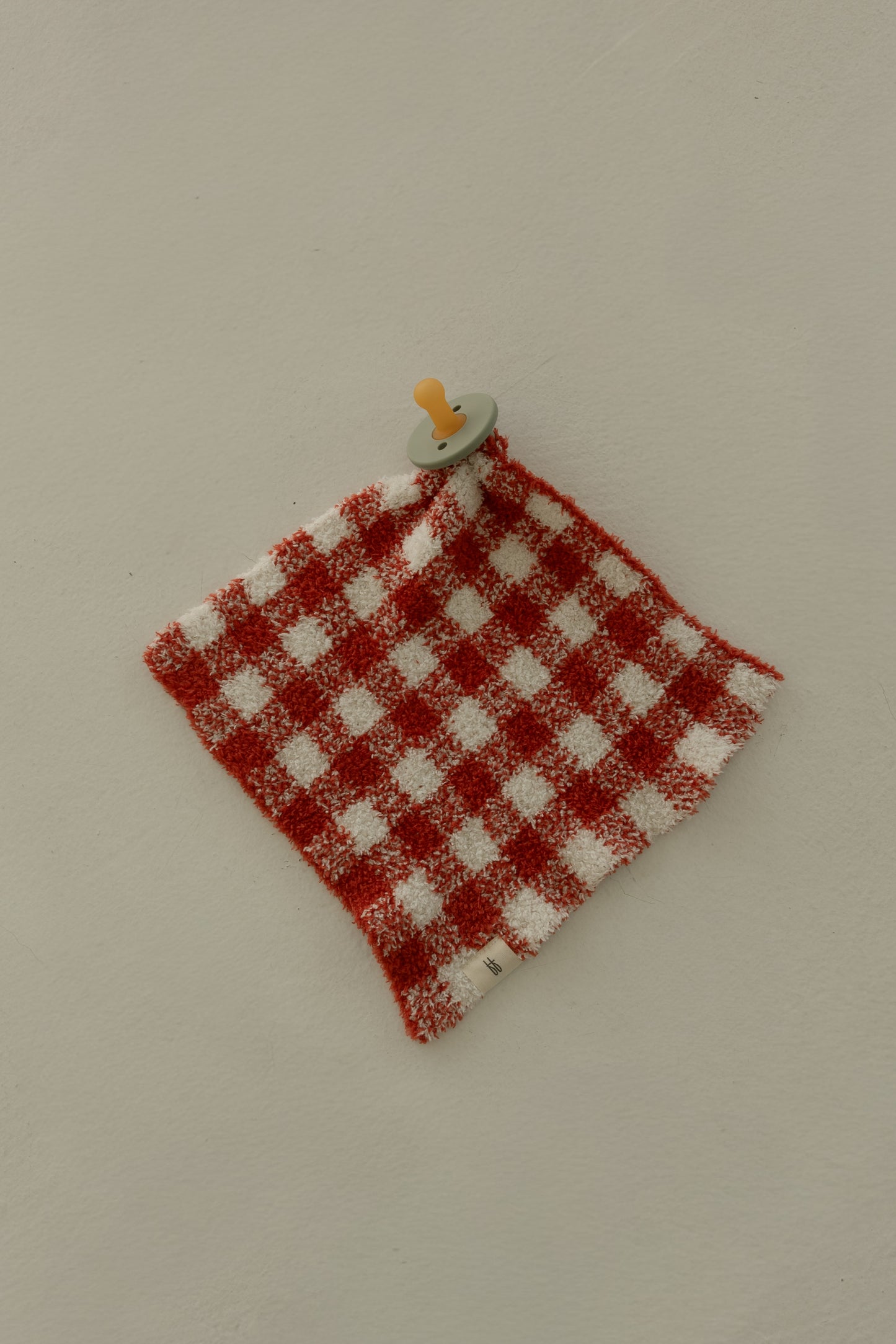 A pacifier is attached to the Lovey | Red Gingham by forever french baby, shown on a light beige surface.