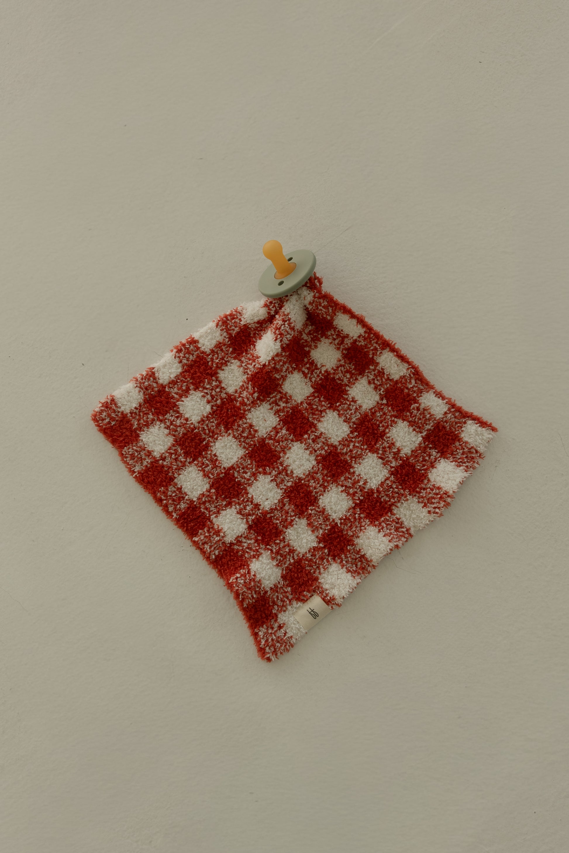 A pacifier is attached to the Lovey | Red Gingham by forever french baby, shown on a light beige surface.