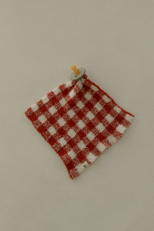 A pacifier is attached to the Lovey | Red Gingham by forever french baby, shown on a light beige surface.