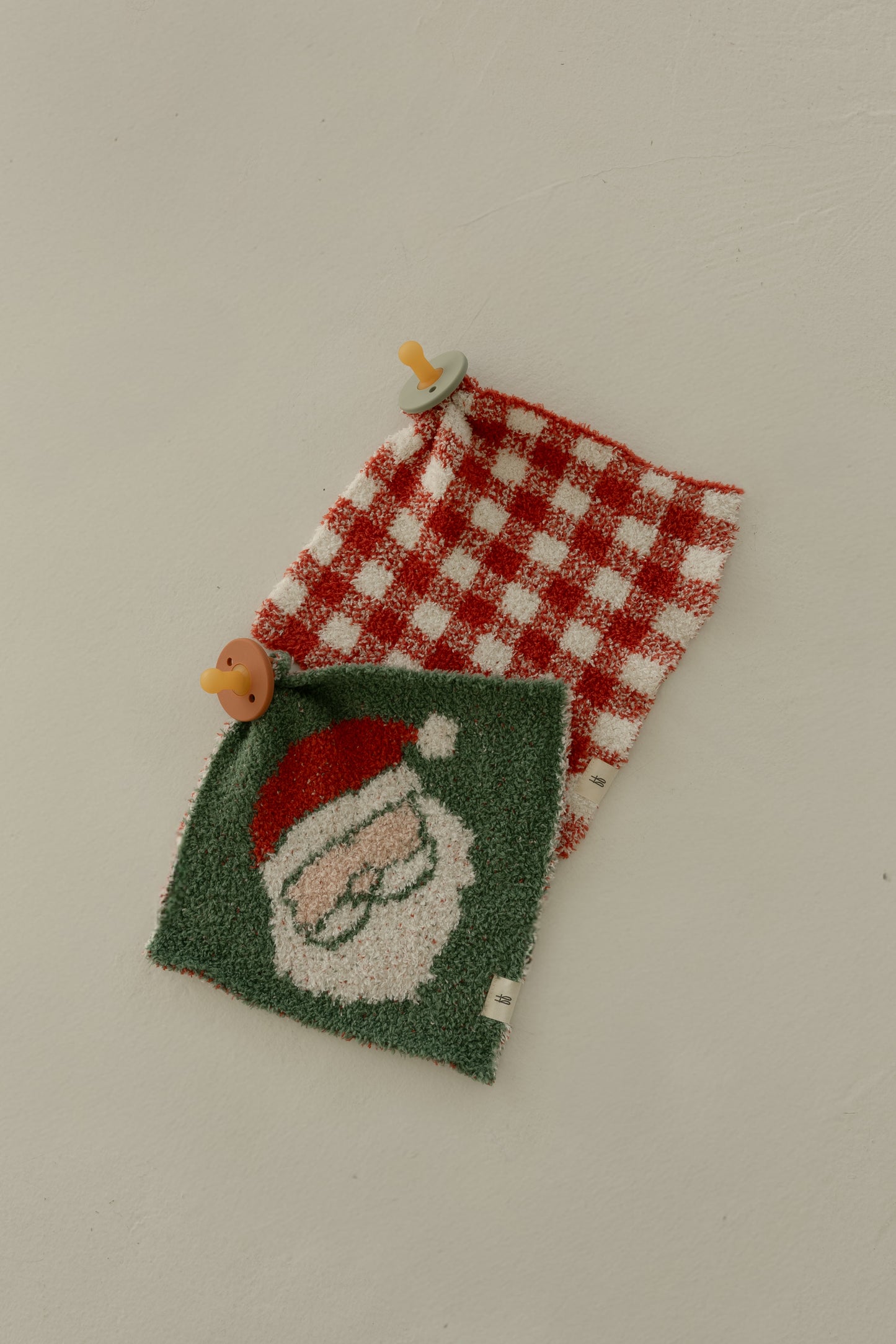 Two colorful pacifiers sit on the Lovey | Santa Checkerboard by forever french baby—a plush cloth with red-and-white checks and a Santa face—displayed atop a soft beige blanket, all set against a light beige background.