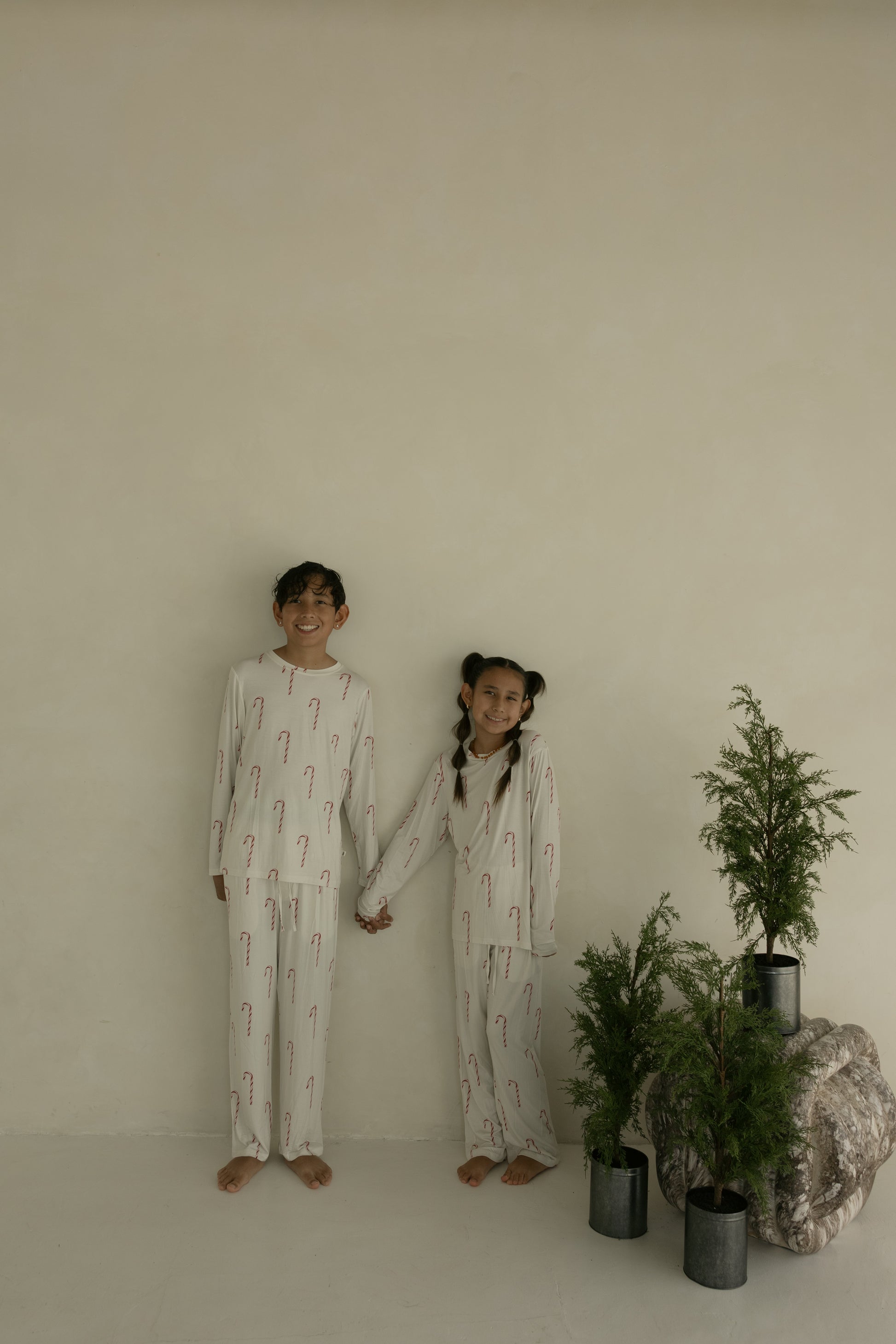 Two children smile and hold hands, barefoot beside potted evergreens, dressed in matching forever french baby Pre-Teen Straight Leg Bamboo Pajamas in the Candy Cane print against a plain, light-colored wall.