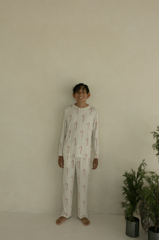 A smiling boy stands barefoot by a beige wall, wearing forever french baby’s Pre-Teen Straight Leg Bamboo Pajamas in the Candy Cane print. Green potted plants to his right add freshness to the cozy bamboo outfit.