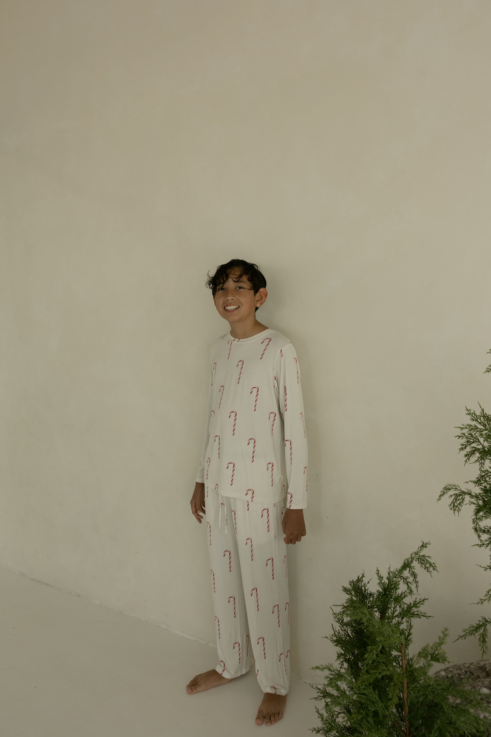 A boy wears forever french baby Pre-Teen Straight Leg Bamboo Pajamas in the Candy Cane pattern, standing barefoot indoors by a green plant and smiling against a plain beige wall.
