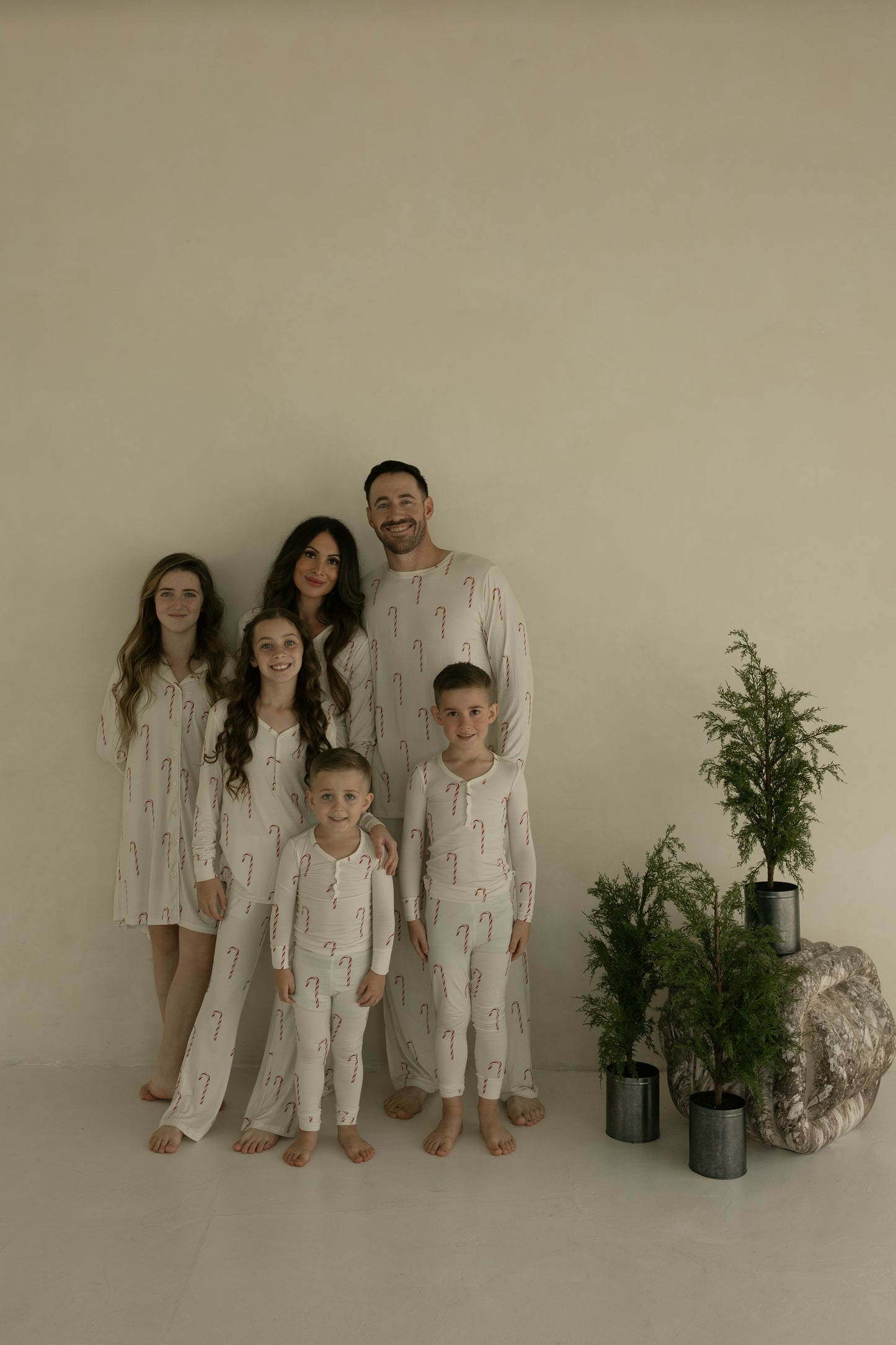 A family of five, all barefoot in matching forever french baby Women's Bamboo Pajamas | Candy Cane, stands indoors by small potted evergreens against a plain beige wall.