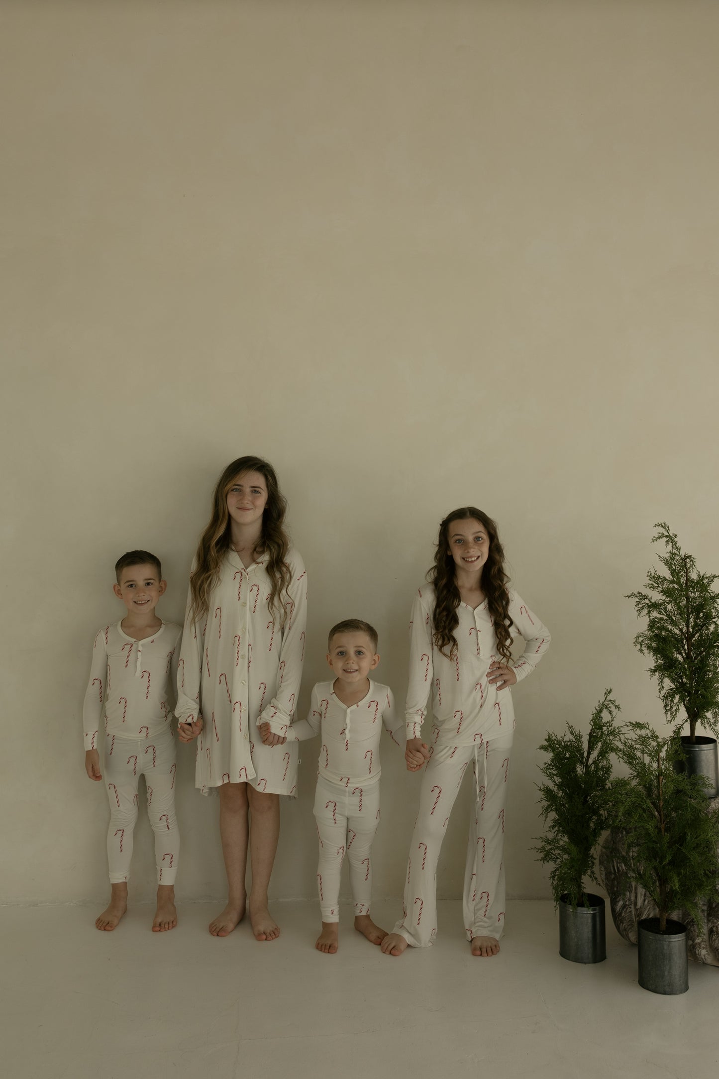 Four children wear matching cream sleepwear with red candy cane prints from forever french baby's Women's Bamboo Sleeping Dress collection, standing barefoot against a beige wall. Small potted evergreens sit on the right.