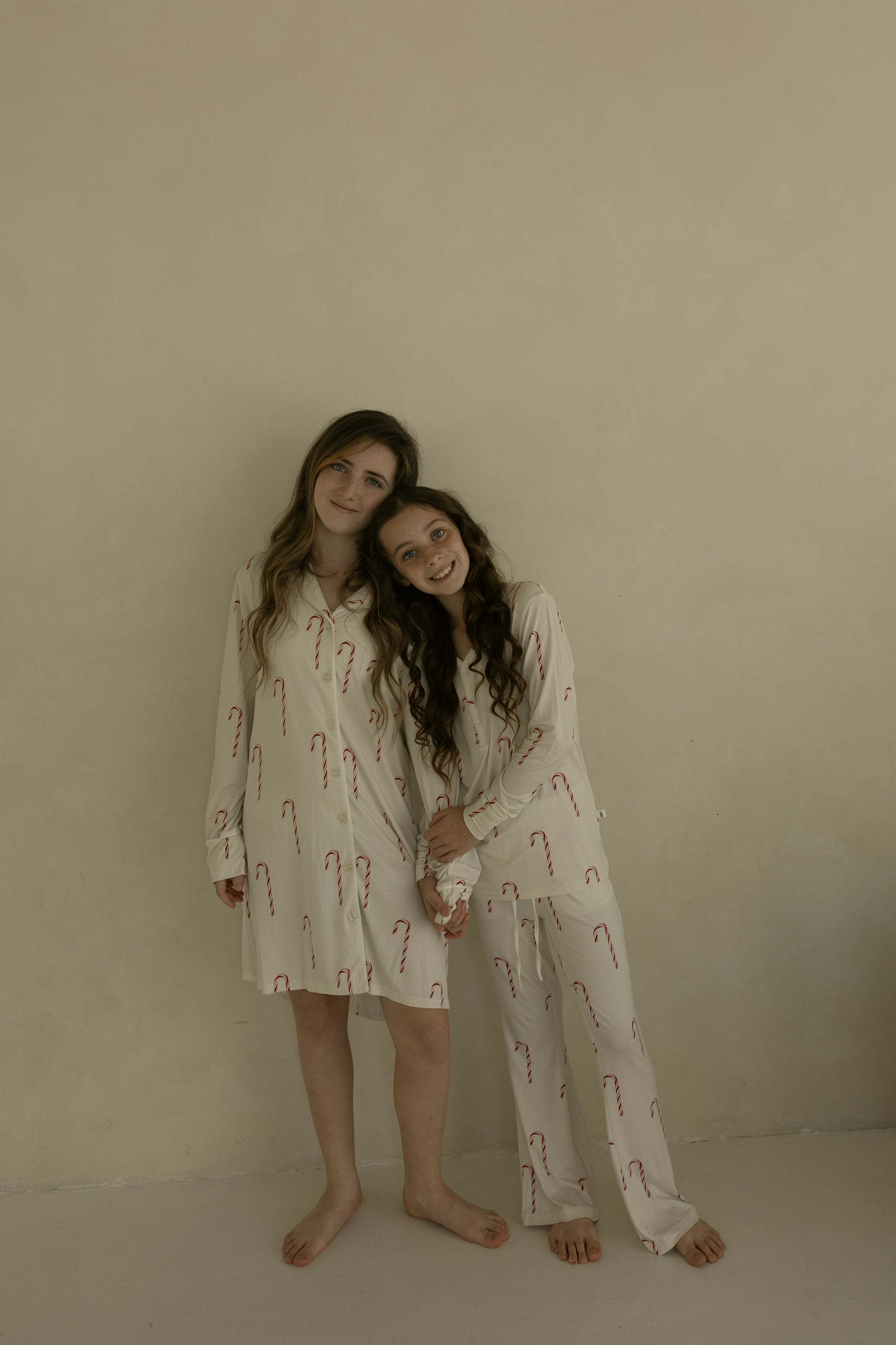 Two girls in matching forever french baby Women's Bamboo Sleeping Dress | Candy Cane stand barefoot against a beige wall, one in the sleeping dress and the other in pajama pants, both smiling and leaning their heads together.