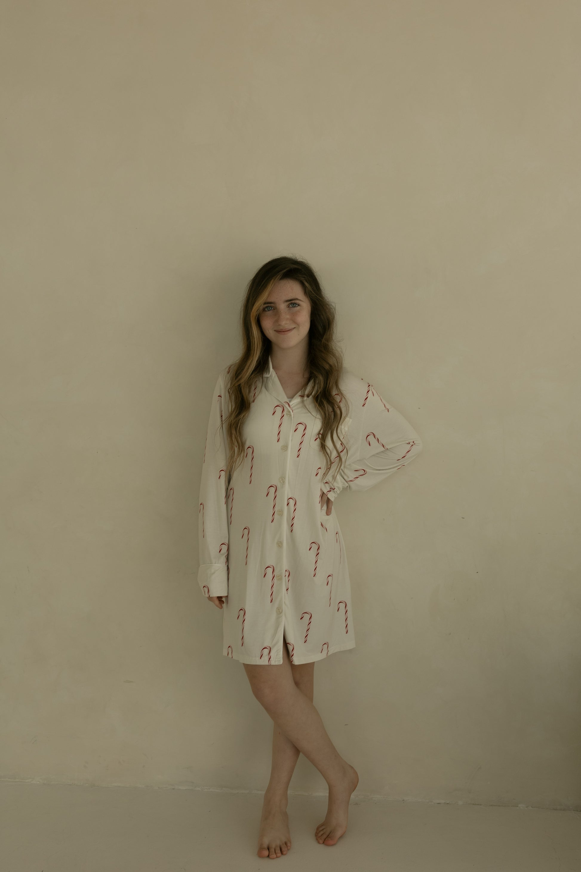 A young woman with long, wavy hair stands barefoot by a beige wall, smiling slightly with one hand on her hip, wearing the forever french baby Women's Bamboo Sleeping Dress | Candy Cane.