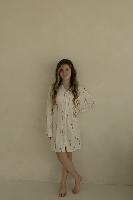 A young woman with long, wavy hair stands barefoot by a beige wall, smiling slightly with one hand on her hip, wearing the forever french baby Women's Bamboo Sleeping Dress | Candy Cane.