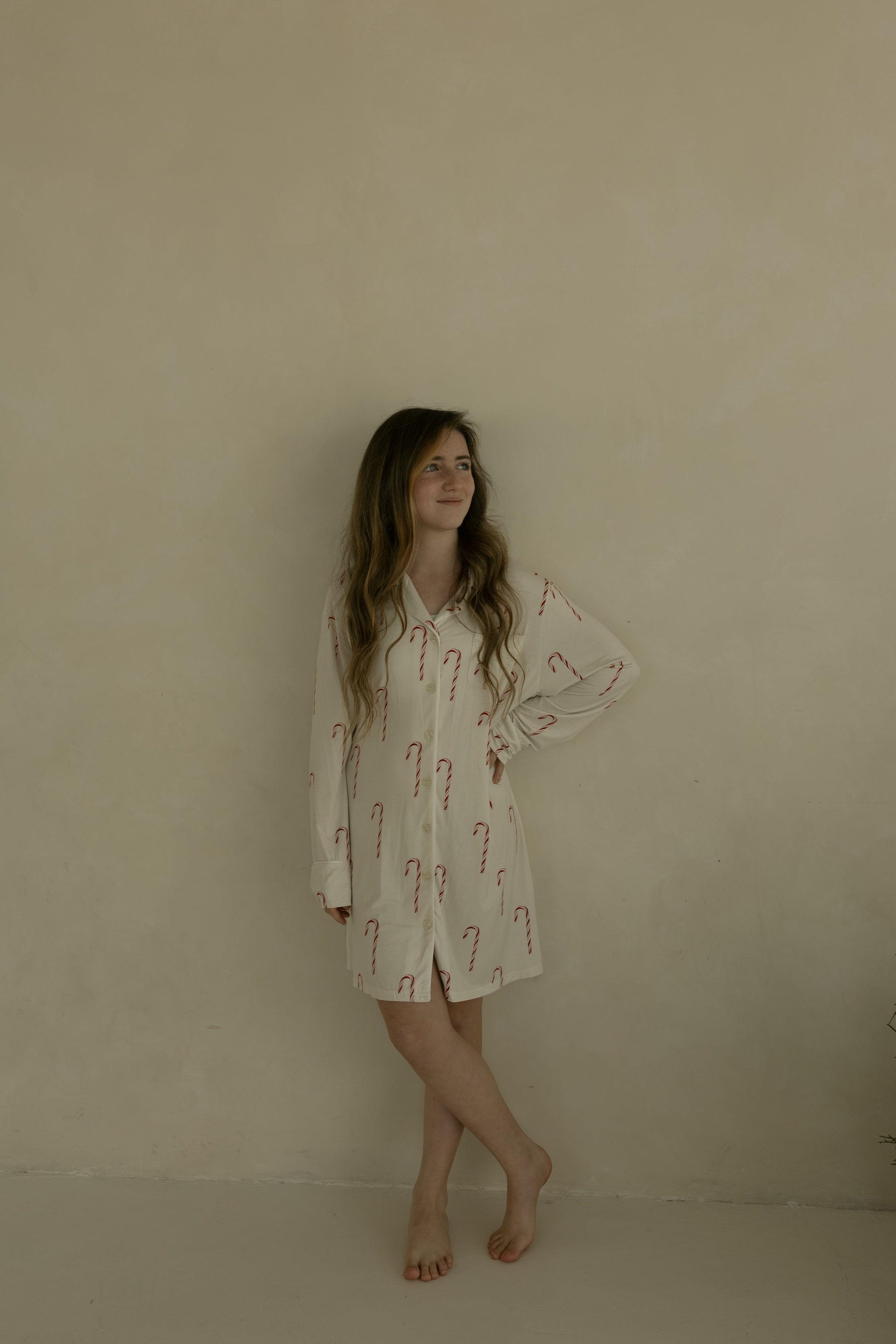 A young woman with long hair stands barefoot against a beige wall, wearing the forever french baby Women's Bamboo Sleeping Dress | Candy Cane. She turns her head slightly to the side, resting one hand on her hip.