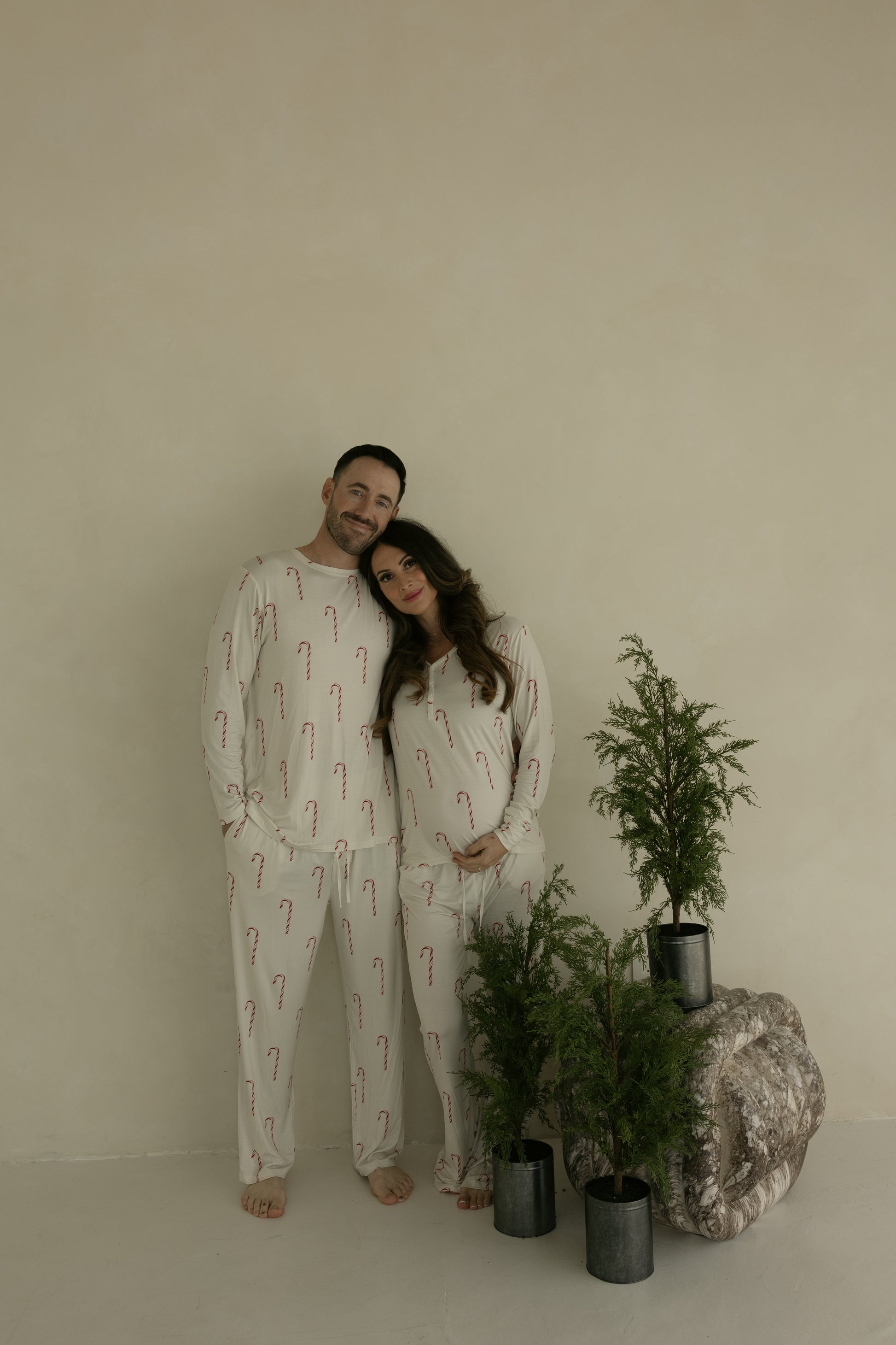 A smiling couple stands barefoot indoors, the man wearing forever french baby's Men's Bamboo Pajamas | Candy Cane. The woman cradles her baby bump beside three small potted evergreens against a plain light wall.