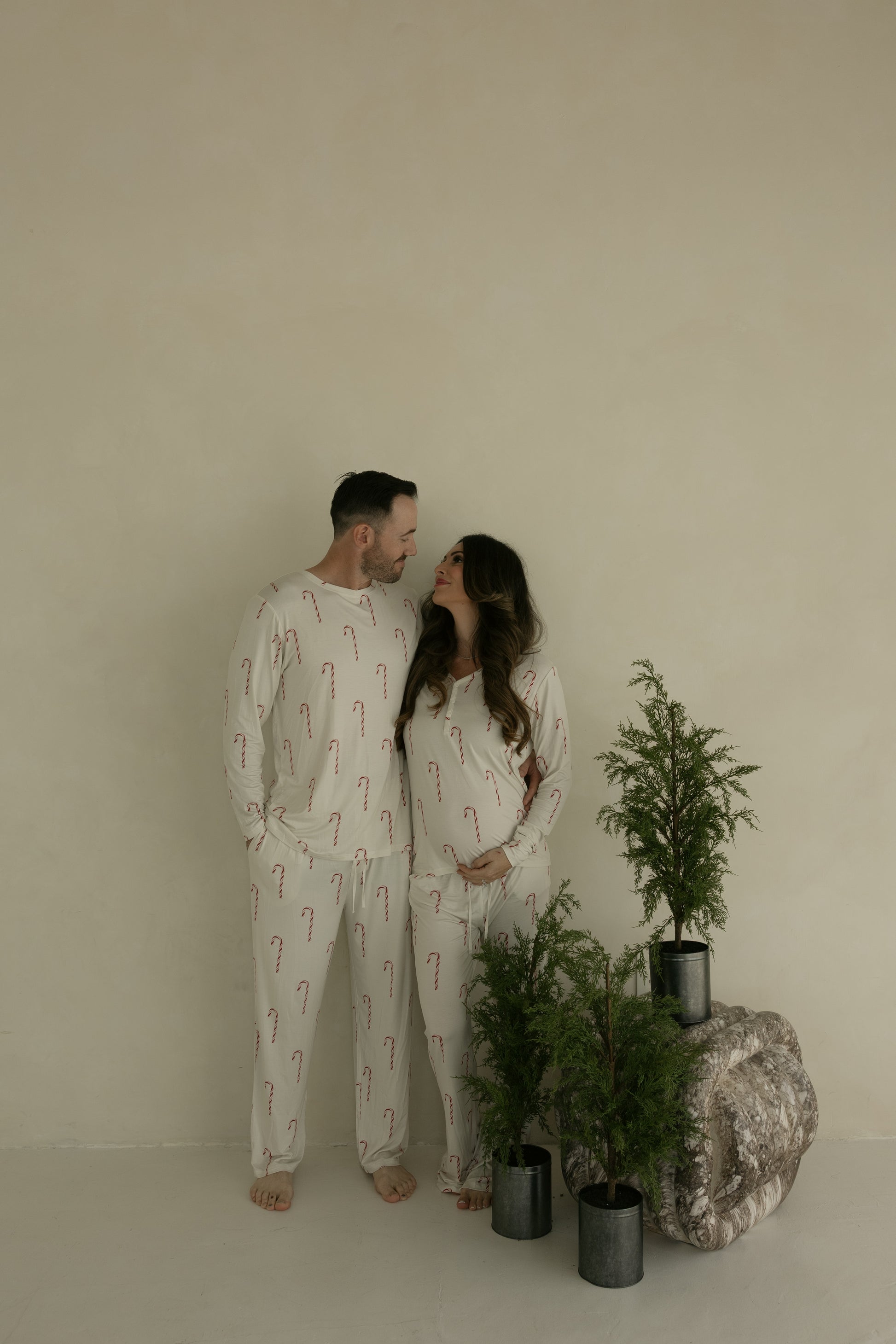 A couple stands barefoot by potted evergreens, wearing matching forever french baby Women's Bamboo Pajamas | Candy Cane. The woman rests her hand on her baby bump. These hypo-allergenic pajamas are cream with red candy canes, set against a neutral background.