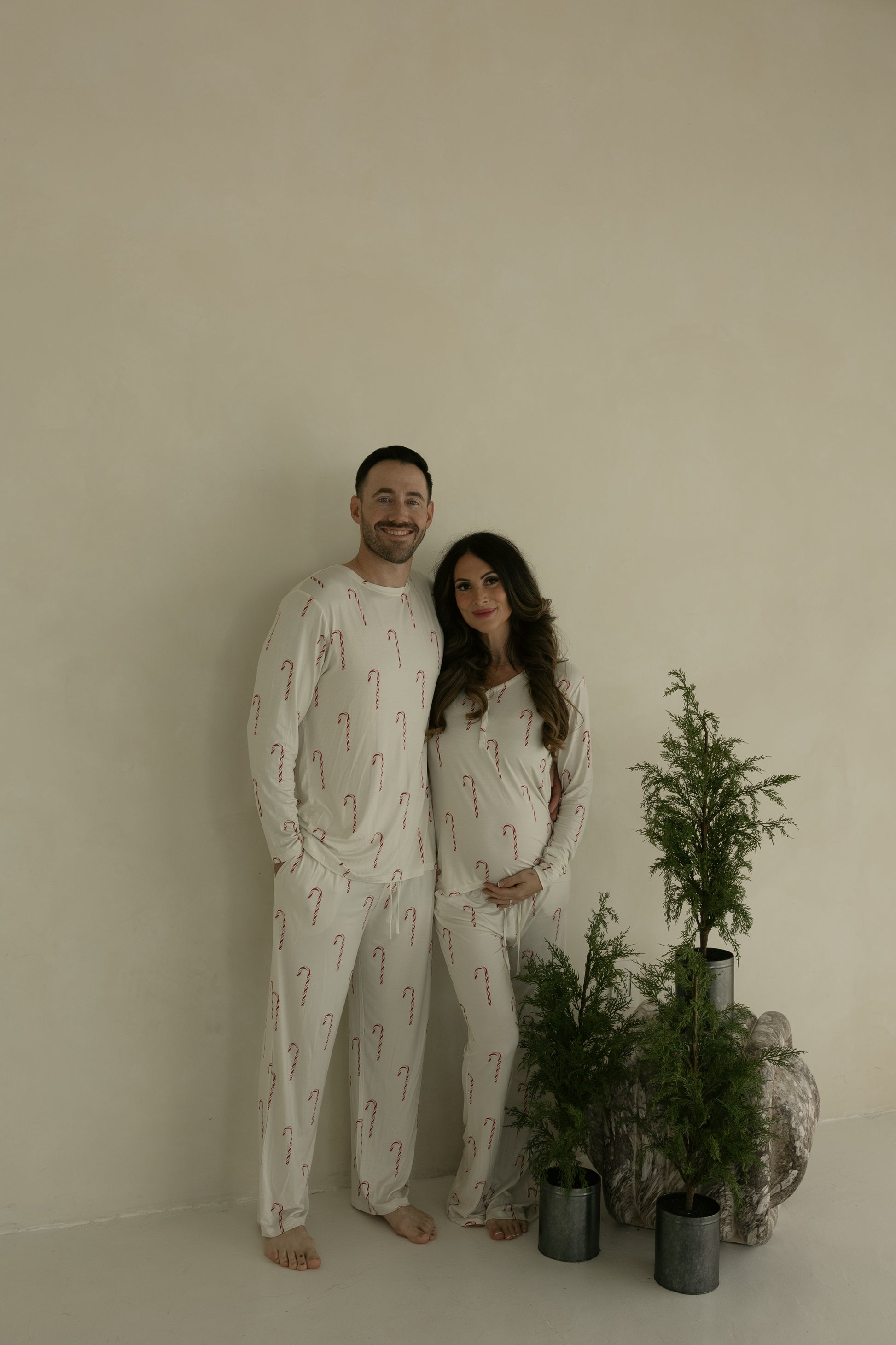 A smiling couple stands barefoot indoors next to potted evergreens; the pregnant woman wears forever french baby Women's Bamboo Pajamas | Candy Cane in cream with red candy canes, resting her hand on her belly.