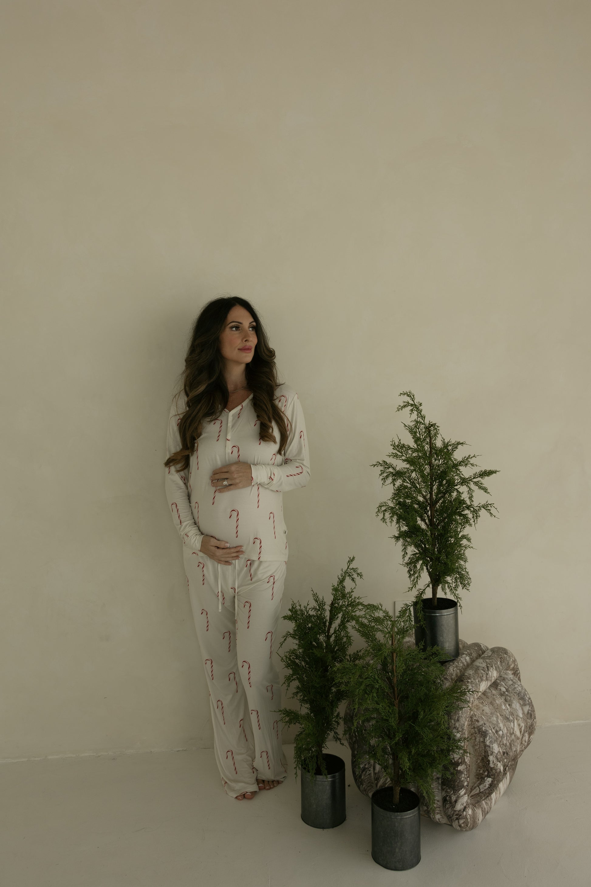A pregnant woman wears forever french baby Women's Bamboo Pajamas | Candy Cane, standing by a light wall and holding her belly, with three small potted evergreen trees on a stone slab nearby.