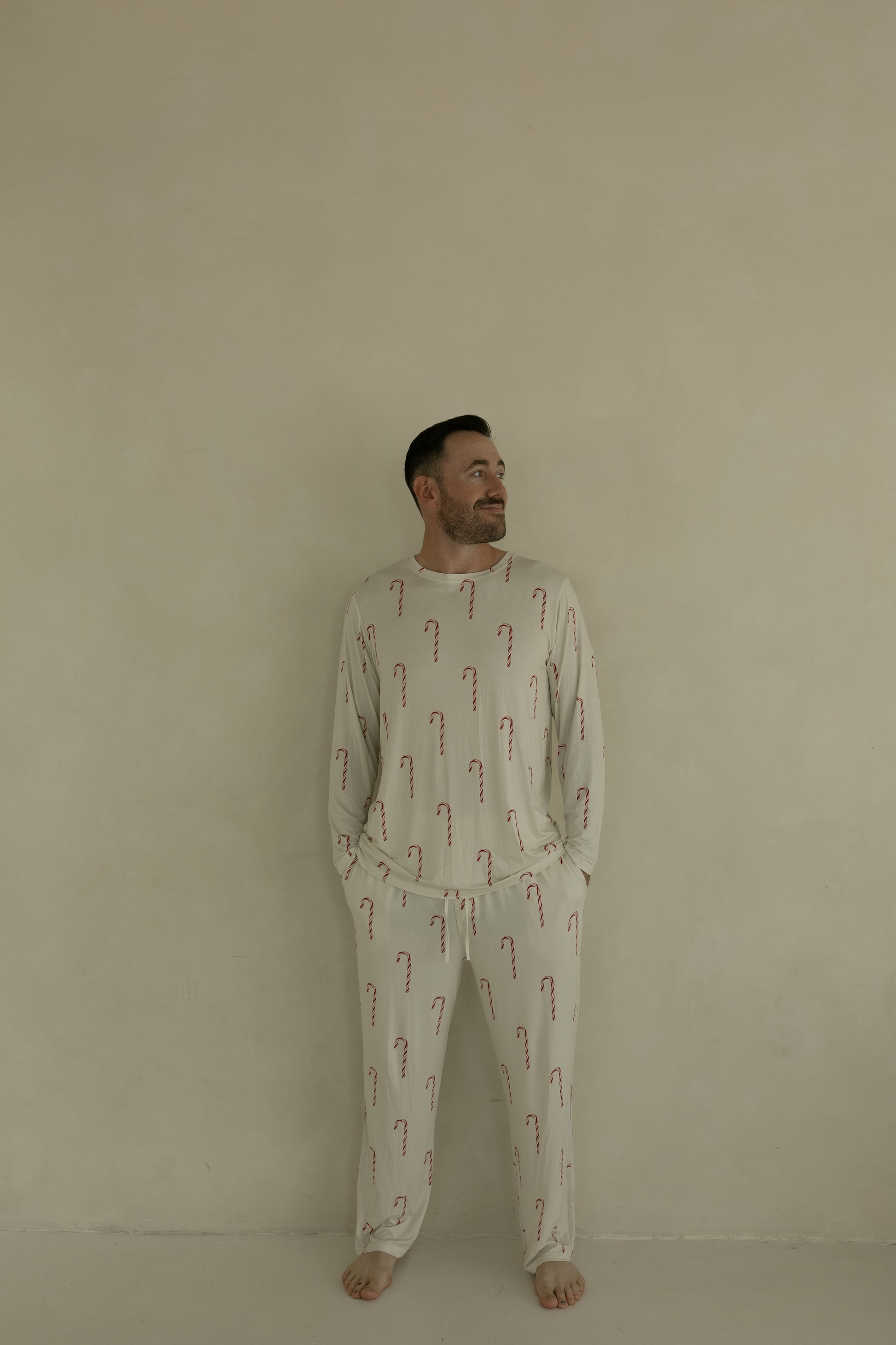 A man stands barefoot against a plain beige wall, wearing forever french baby Men's Bamboo Pajamas | Candy Cane, featuring a light-colored fabric with red candy cane pattern. He has his hands in his pockets and is smiling slightly while looking to the side.