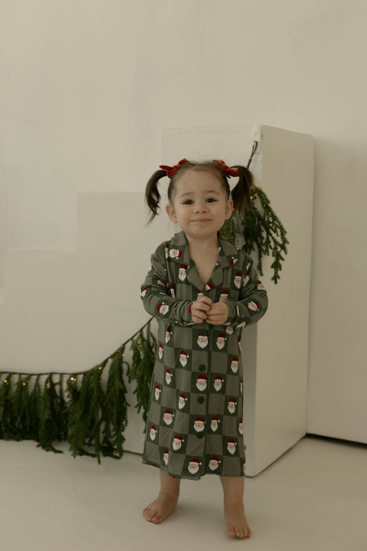 A barefoot young child with pigtails and red bows smiles indoors, wearing the forever french baby Toddler Bamboo Sleeping Dress in Santa Checkerboard, surrounded by greenery and string lights against a white background.