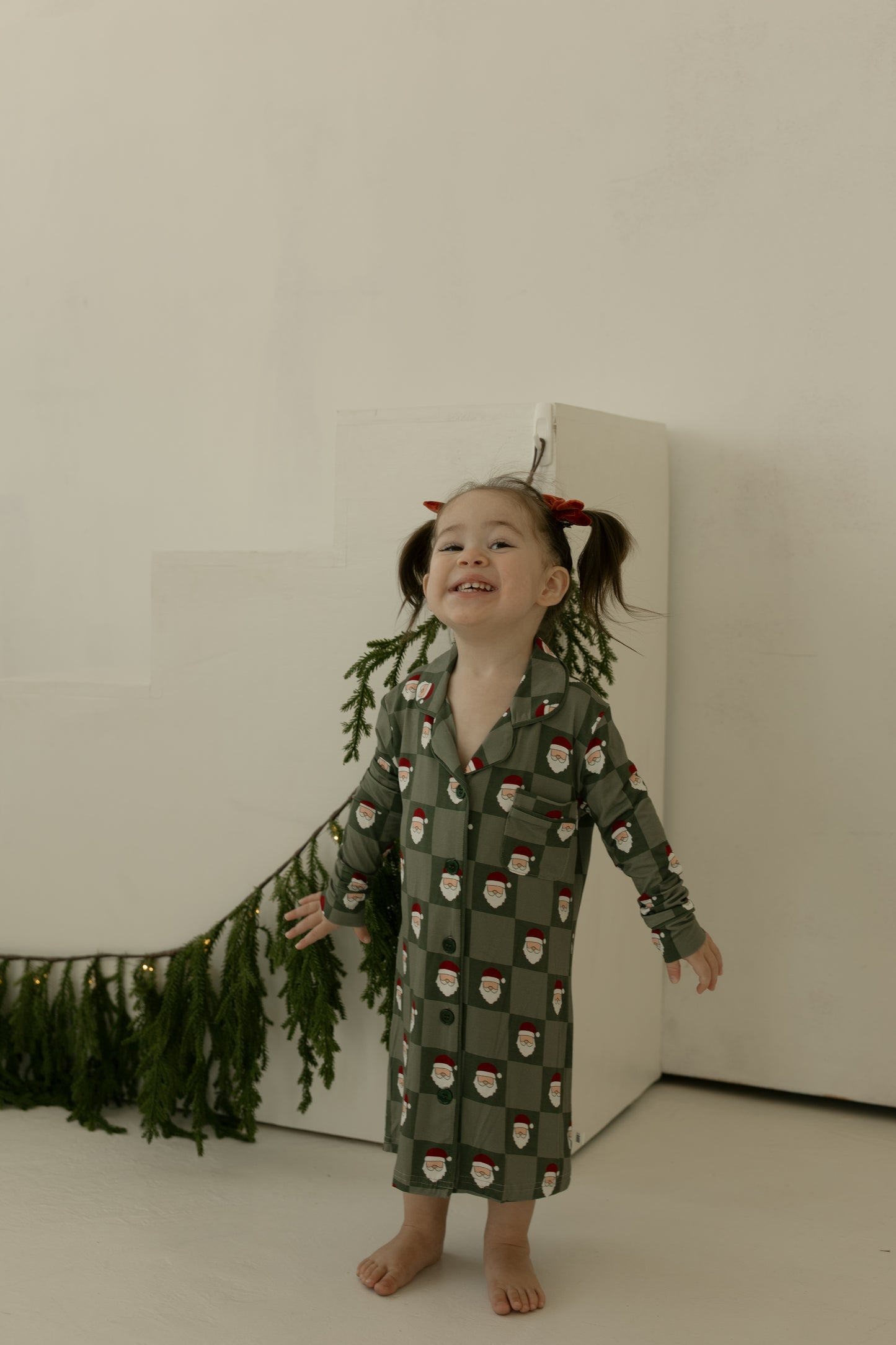 A young girl with pigtails and red bows smiles barefoot on white stairs, wearing the forever french baby Toddler Bamboo Sleeping Dress in Santa Checkerboard. Holiday greenery lines the steps.