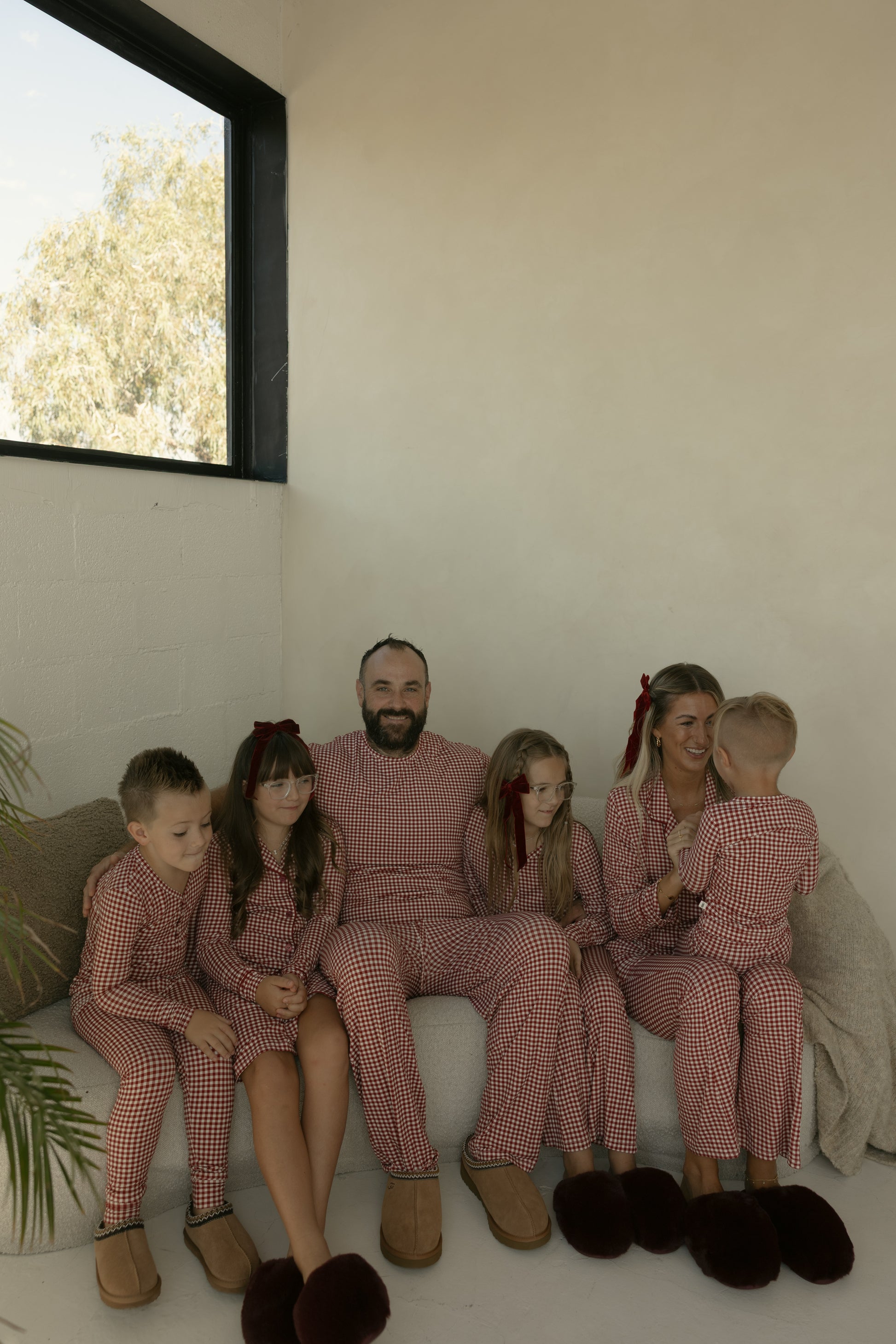 A family of five, all dressed in forever french baby Bamboo Two Piece Pajamas in Red Gingham, smiles together on a sofa in a softly lit room.