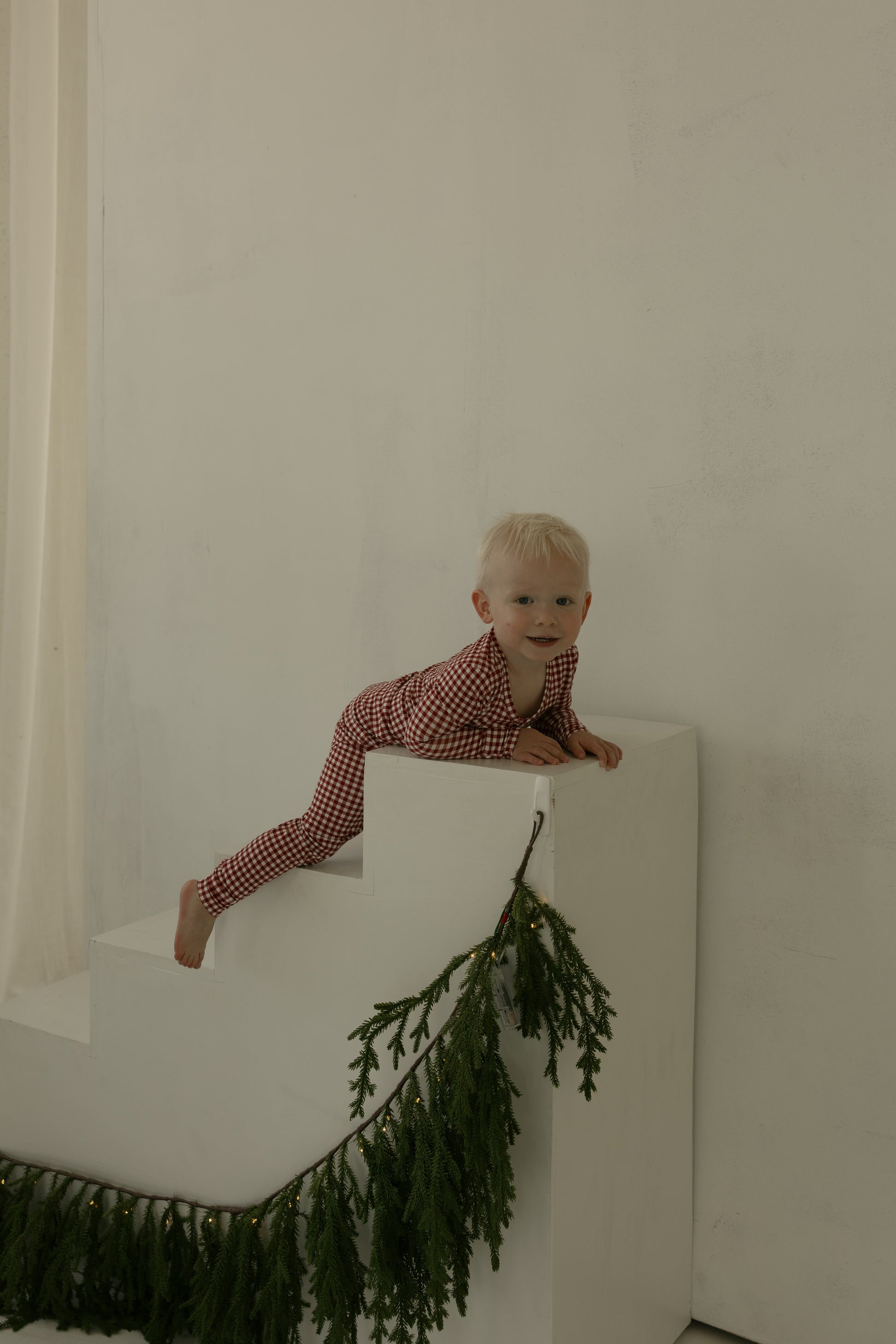 A young child in forever french baby's Bamboo Two Piece Pajamas | Red Gingham climbs white steps with pine garland and lights, against a plain background—showcasing stylish, hypoallergenic sleepwear for cozy nights.