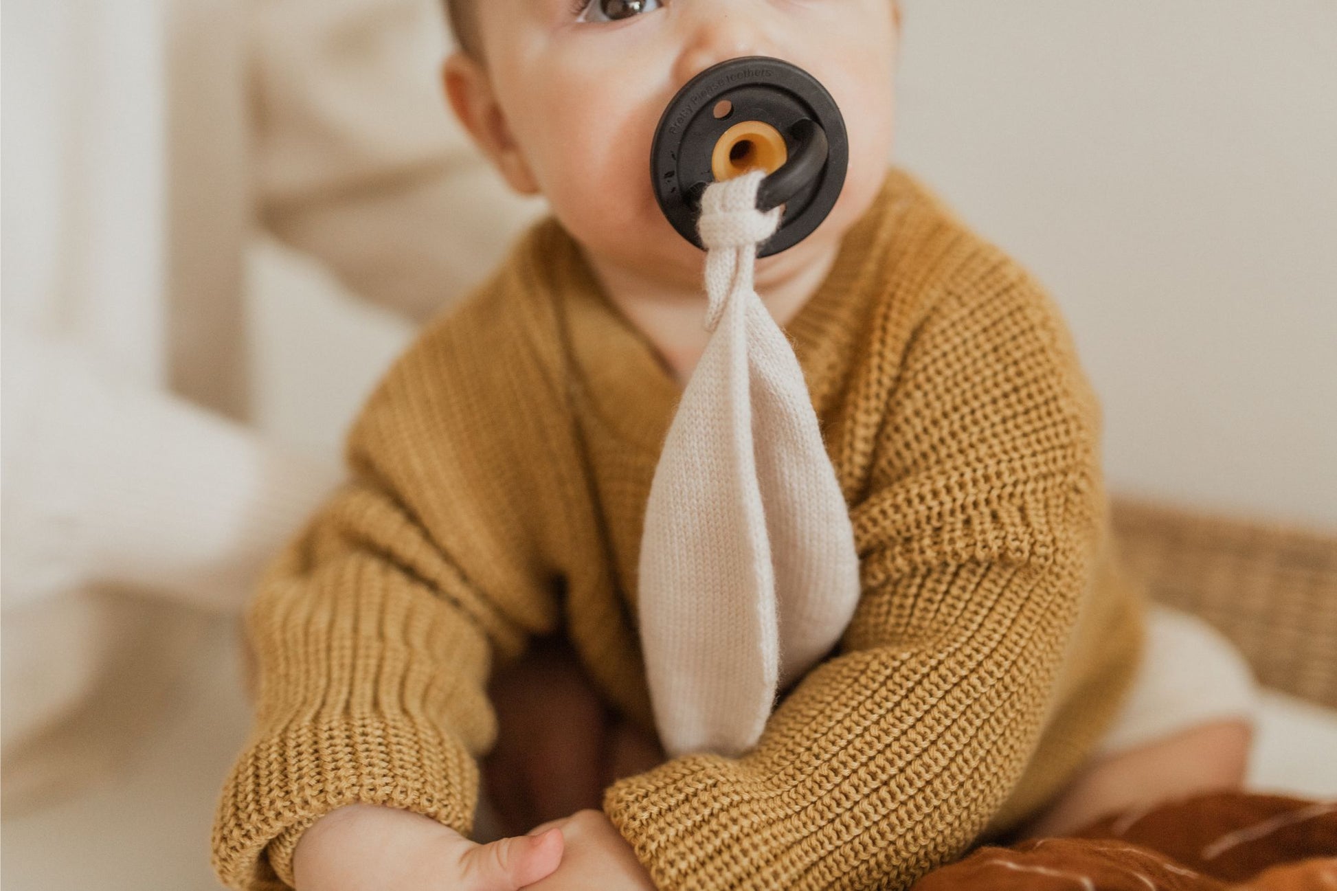 A baby in a mustard-colored knitted sweater is sucking on the Modern Pacifier in Warm Taupe by forever french baby, which has a cloth attached to it. The BPA-free, silicone design ensures safety and comfort as the baby sits holding their foot. The softly blurred background suggests an indoor setting.