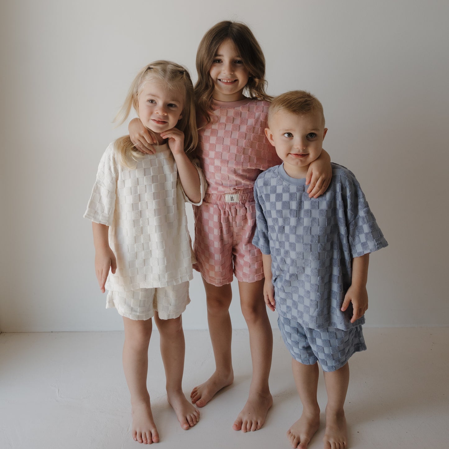 Three young children stand barefoot against a white background, smiling in family matching outfits. The boy on the right wears the forever french baby Child Terry Short Set | Tradewinds.