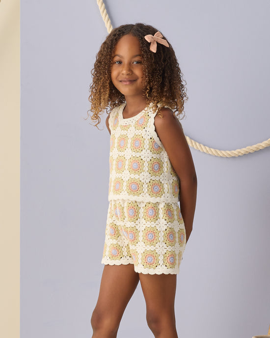 A young girl with curly hair and a pink bow smiles, wearing the Rylee + Cru Crochet Tank Set in Citron Daisy Check—a sleeveless outfit with delicate ruffled trim and matching shorts. The background is light purple with a thick rope arranged in a loop.