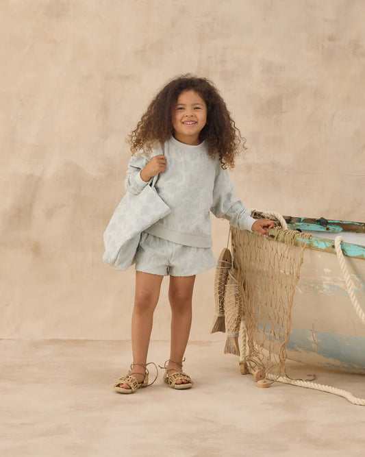 A young child with curly hair wears the Rylee + Cru Karli Set in soft terry cloth—a light sweater and shorts ensemble—standing beside a weathered wooden boat draped in netting, creating a relaxed coastal vibe against a soft beige backdrop.