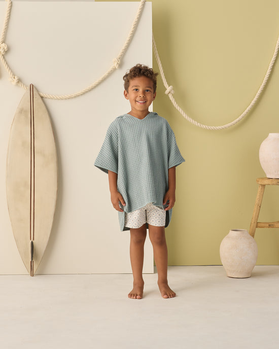 A young child stands barefoot indoors next to a surfboard, pottery, and wooden stool against a backdrop decorated with thick, draped rope. They are wearing Rylee + Cru’s Boardshort | Green Fleur—UPF 50+ swimwear made from recycled polyester.