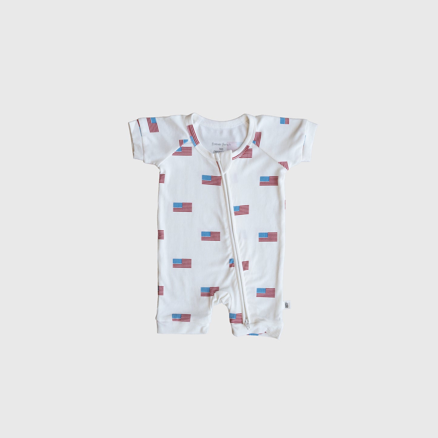 The forever french baby Shortie Bamboo Zip | Americana Ribbed is a white, short-sleeve romper with a zipper front and repeating American flags on light gray—stylish, hypoallergenic babywear for patriotic style.