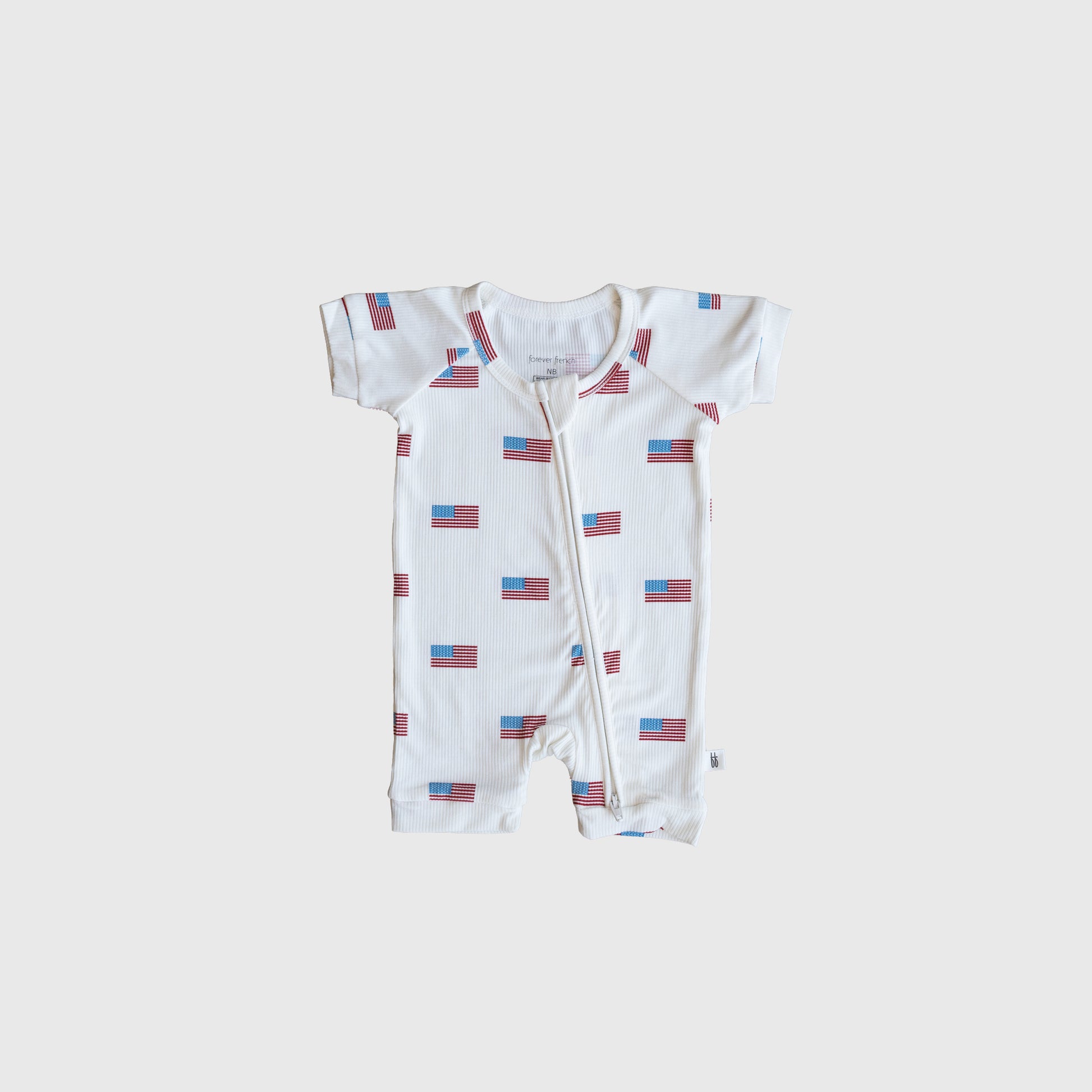 The forever french baby Shortie Bamboo Zip | Americana Ribbed is a white, short-sleeve romper with a zipper front and repeating American flags on light gray—stylish, hypoallergenic babywear for patriotic style.