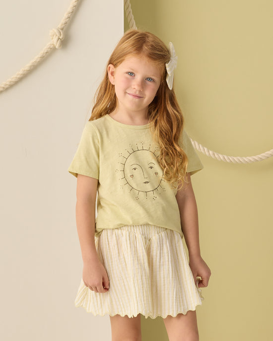 A young girl with long red hair and a white bow wears a yellow sun-face graphic tee paired with the Smocked V Mini Skirt in Citron Stripe by Rylee + Cru, featuring a light pleated design and delicate scalloped hem. She stands against a beige and green wall adorned with rope decoration.