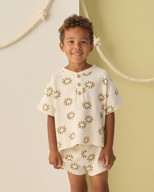 A young child with short curly hair smiles, wearing the Rylee + Cru Summer Waffle Set | Sunny—featuring a matching cream-colored shirt and 100% cotton waffle fabric shorts with a brown sunny print—while standing against a two-tone background accented with decorative rope.