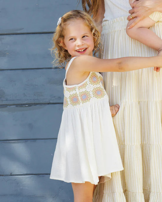A smiling young girl stands outside in front of a blue wooden wall, holding a woman’s arm. She wears the Rylee + Cru Crochet Sundress in Citron Daisy Check, featuring a delicate crochet bodice and white dress. The woman beside her, dressed in cream, is partly visible.
