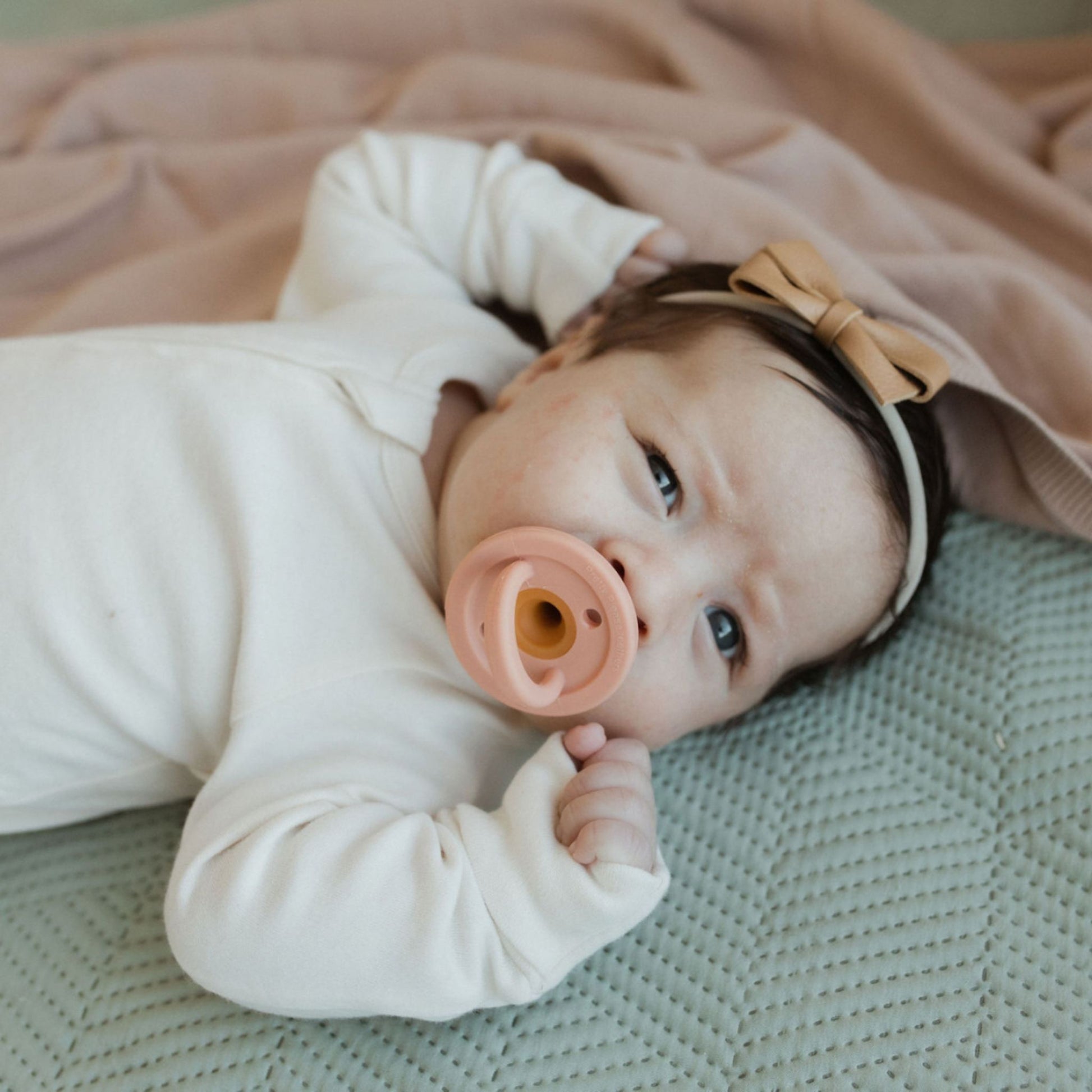 A baby lies on a textured green blanket, looking up with a curious expression, wearing a cream-colored onesie and a peach-colored bow headband. The baby has one of those Modern Pacifiers in Caramel from forever french baby in their mouth and is partially covered by a pink blanket.