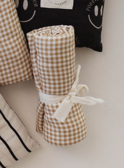 A rolled-up *Golden Gingham* muslin swaddle from **forever french baby**, tied with a white string, is placed on a light surface. Surrounding it are parts of other fabric items, including a black piece with smiley faces and a striped fabric. The cloth evokes the charm of a vintage baby blanket or muslin swaddle.