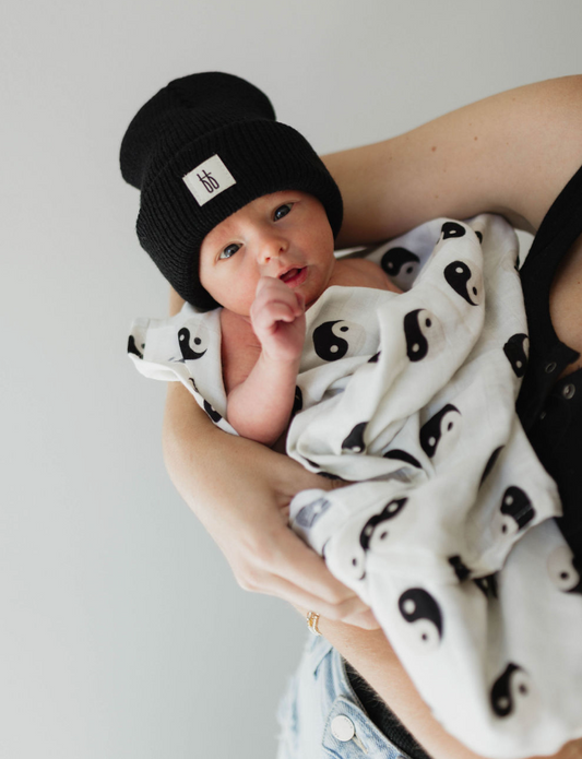 Cradled gently in someone’s arms, a newborn baby is wrapped in the forever french baby’s Muslin Swaddle | Black & White Yin Yang (misprint), with a cozy black beanie completing the look.