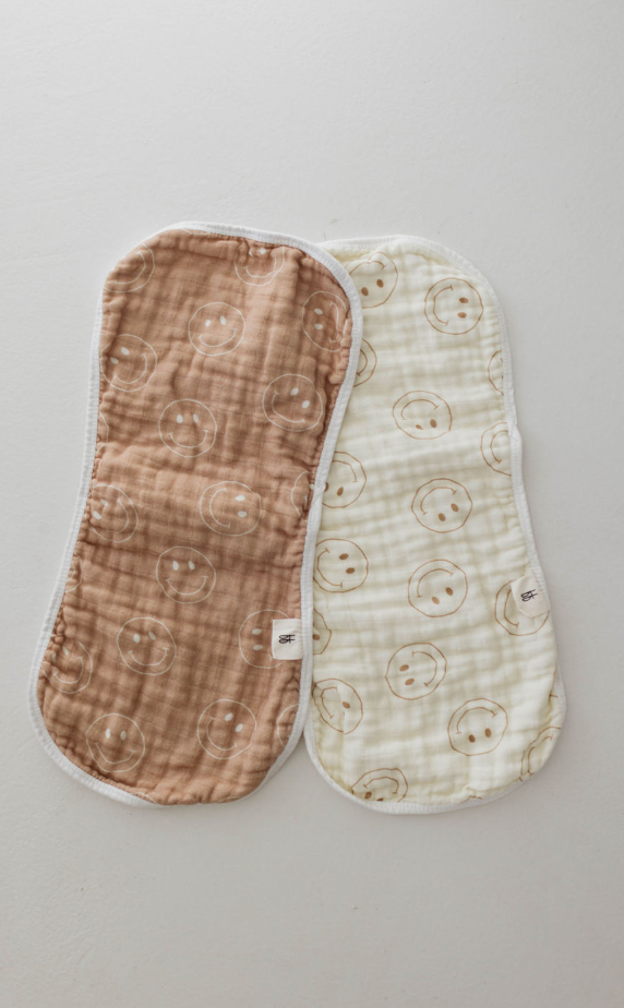 Two "Just Smile Tan" 100% cotton burp cloths from forever french baby are shown on a light surface. The tan burp cloth on the left features a smiley face pattern, while the ivory one on the right boasts a similar design. Both have white edges and textured fabric.
