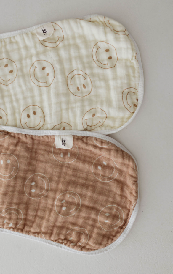 Two oval-shaped quilted burp cloths from forever french baby, part of the "Just Smile Tan" collection, feature a pattern of simple smiley faces. One is ivory-colored and the other brown. The ivory burp cloth is positioned above the brown one on a light grey surface, and both are made from 100% cotton.