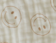 The Burp Cloth | Just Smile Tan from forever french baby is a light beige, 100% cotton fabric featuring a pattern of brown smiley faces, some right-side up and others upside down. This tan burp cloth appears soft and airy, with subtle creases.