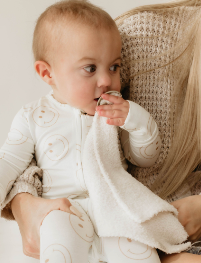 A baby in a white outfit decorated with smiley faces chews on the soft microfibre feather yarn "Retro Christmas" lovey from forever french baby while sitting on a woman's lap. The woman, wearing a textured beige sweater, adds to the warm, cozy atmosphere of the scene.