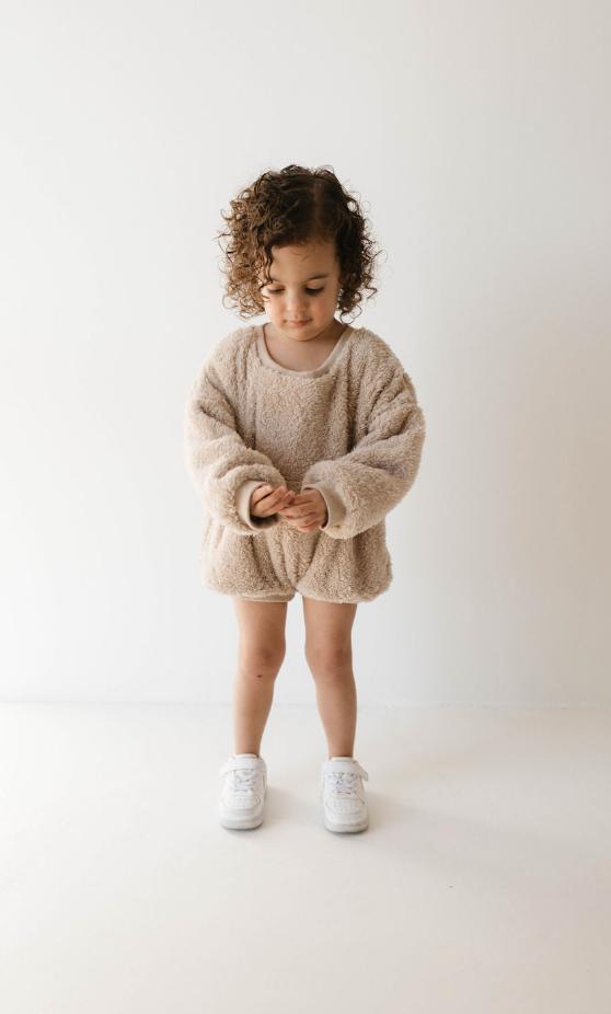 A young child with curly hair stands in front of a plain white background. They are wearing an oversized, beige textured sweater and white sneakers. The child's calm expression shifts slightly as they gaze down at their hands, clad in the cozy fabric of the Long Sleeve Romper | Teddy Bear by forever french baby.