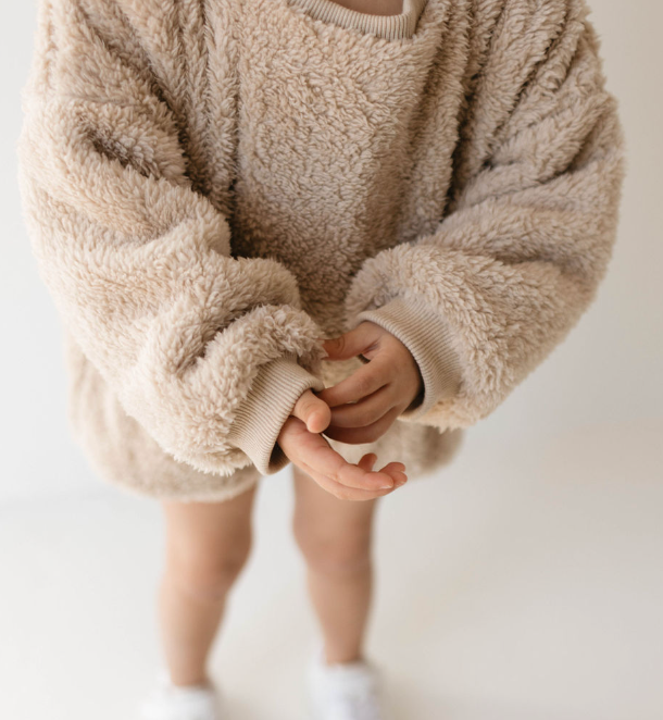 A child dressed in a slightly oversized, fluffy Long Sleeve Romper | Teddy Bear from forever french baby stands with their hands clasped together. The bottom part of their legs is visible, and they are wearing white shoes. The background is white and minimalistic.