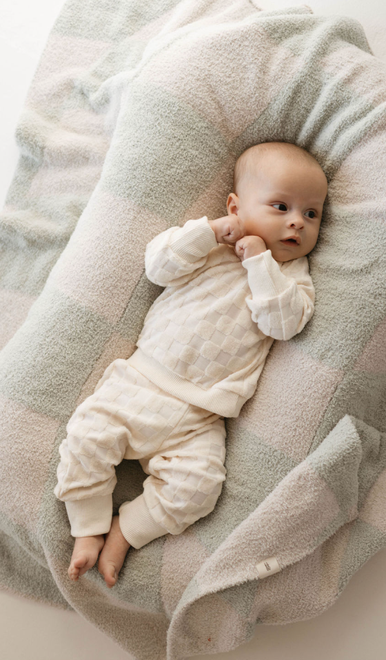 A baby lying on a soft, pastel-colored blanket, dressed in the cozy Long Sleeve Set | Cream Terry Checkerboard by forever french baby. The baby has a calm expression and is looking slightly to the side with arms bent near the face.