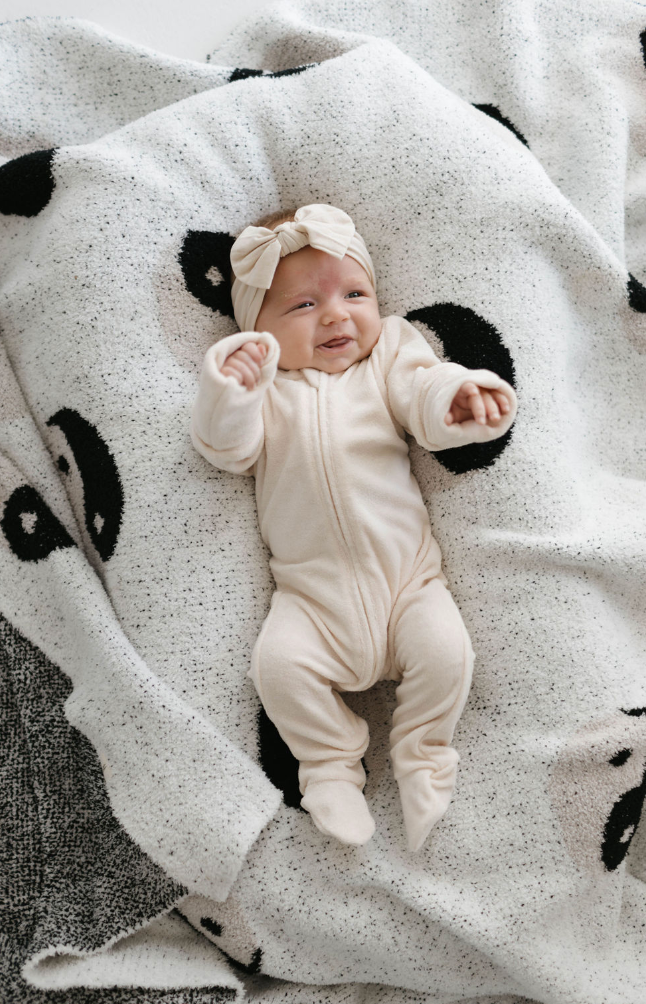 A smiling baby dressed in forever french baby's Vanilla Terry Zip Pajamas and a matching headband lies on a blanket with a black and white panda design. The baby has arms extended upwards, appearing relaxed and happy, enjoying the cozy snuggle feel.