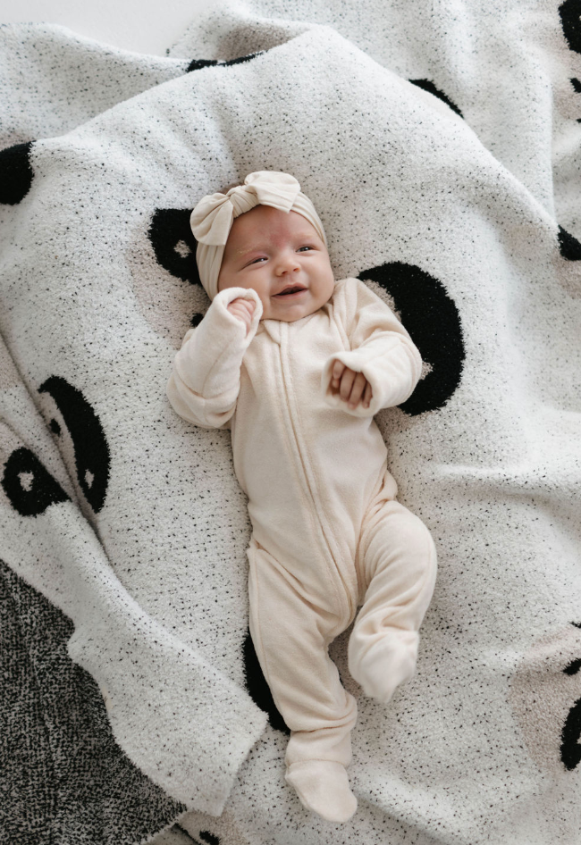 A baby lies on a blanket wearing the Vanilla Terry Zip Pajamas from forever french baby, featuring cozy, cream-colored zip-up footed pajamas and a matching headband with a bow. The baby has a small smile and one hand resting on their cheek. The blanket is white with black circular patterns.