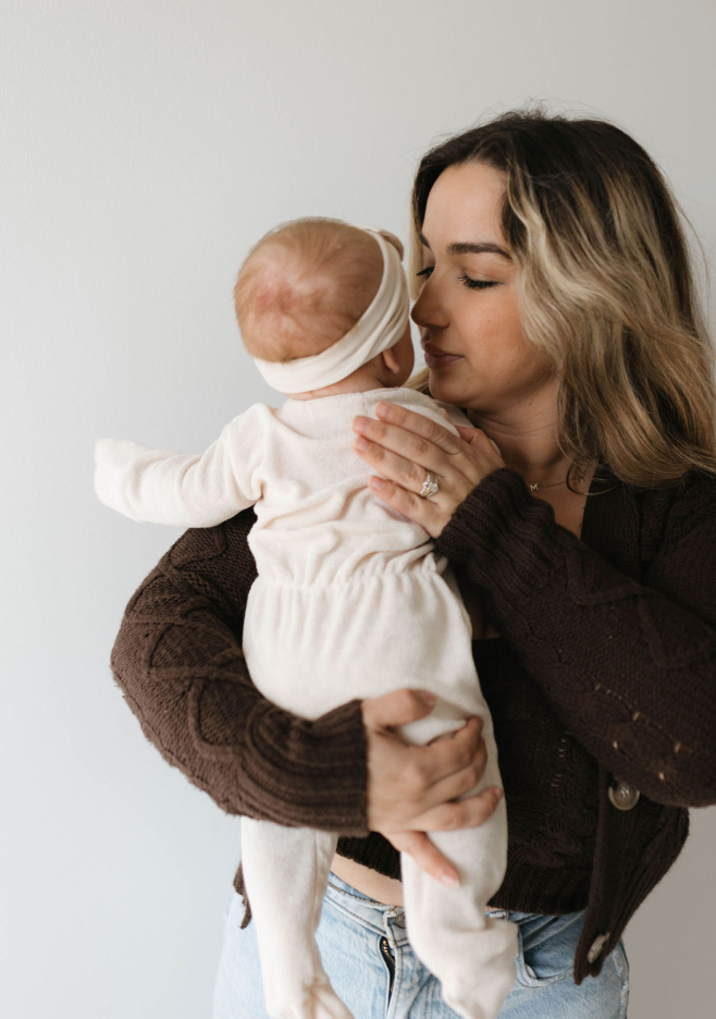 A woman with wavy hair, wearing a dark sweater and light blue jeans, tenderly holds an infant dressed in forever french baby's Vanilla Terry Zip Pajamas. The baby's back is to the camera as the woman touches her nose to the baby's, enjoying a cozy snuggle feel. The background is plain white.