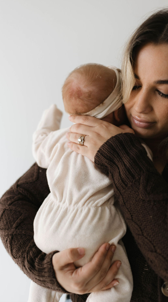 A woman wearing a brown sweater gently cradles a baby dressed in forever french baby's Vanilla Terry Zip Pajamas. The woman's eyes are closed as she holds the baby close, her ring-adorned hand resting tenderly on the baby's back. Both exude a sense of calm and tenderness, wrapped in the cozy snuggle feel of their embrace.