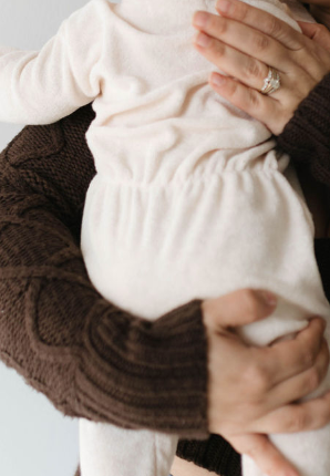 An adult wearing a brown knit sweater holds a baby dressed in cozy Vanilla Terry Zip Pajamas by Forever French Baby. The adult's hands cradle the baby's back and legs, with one hand revealing a diamond ring on the ring finger.