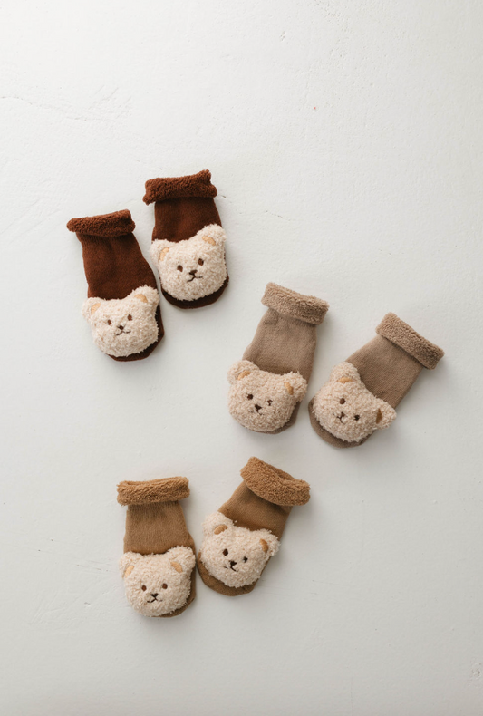 Three pairs of cozy, woolen infant socks from forever french baby are arranged on a light-colored surface. Each pair of the Teddy Bear Grip Socks features an animal face design on the toes: two pairs in beige and tan, and one pair in brown and tan. These socks have a grip on the bottom to prevent slipping.